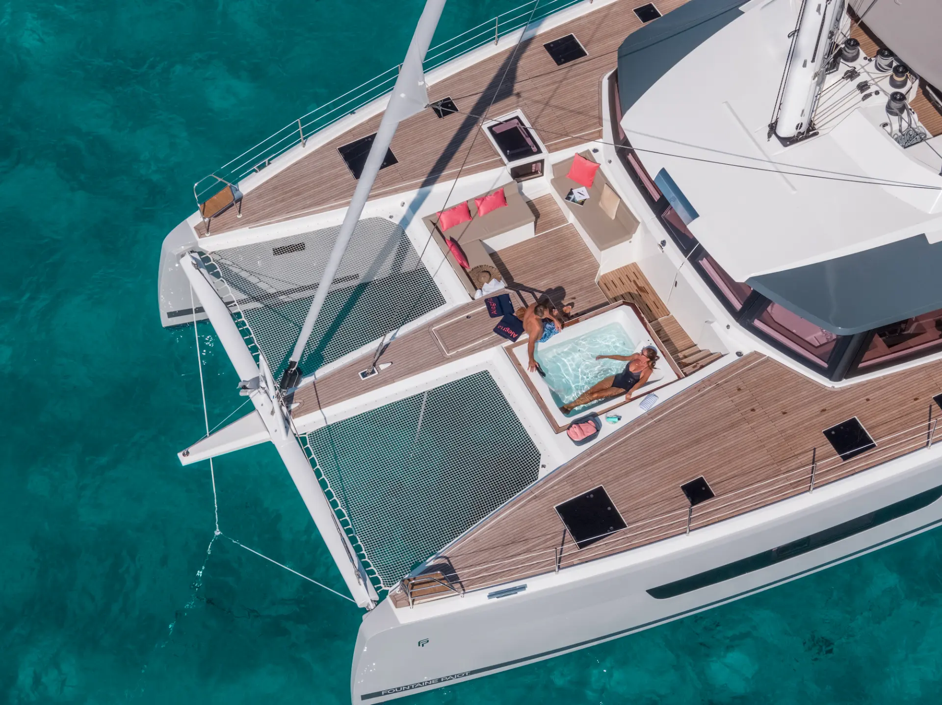 Luxury catamaran Fountaine Pajot 67 - close aerial view of foredeck jacuzzi with lady inside and man outside lounging.