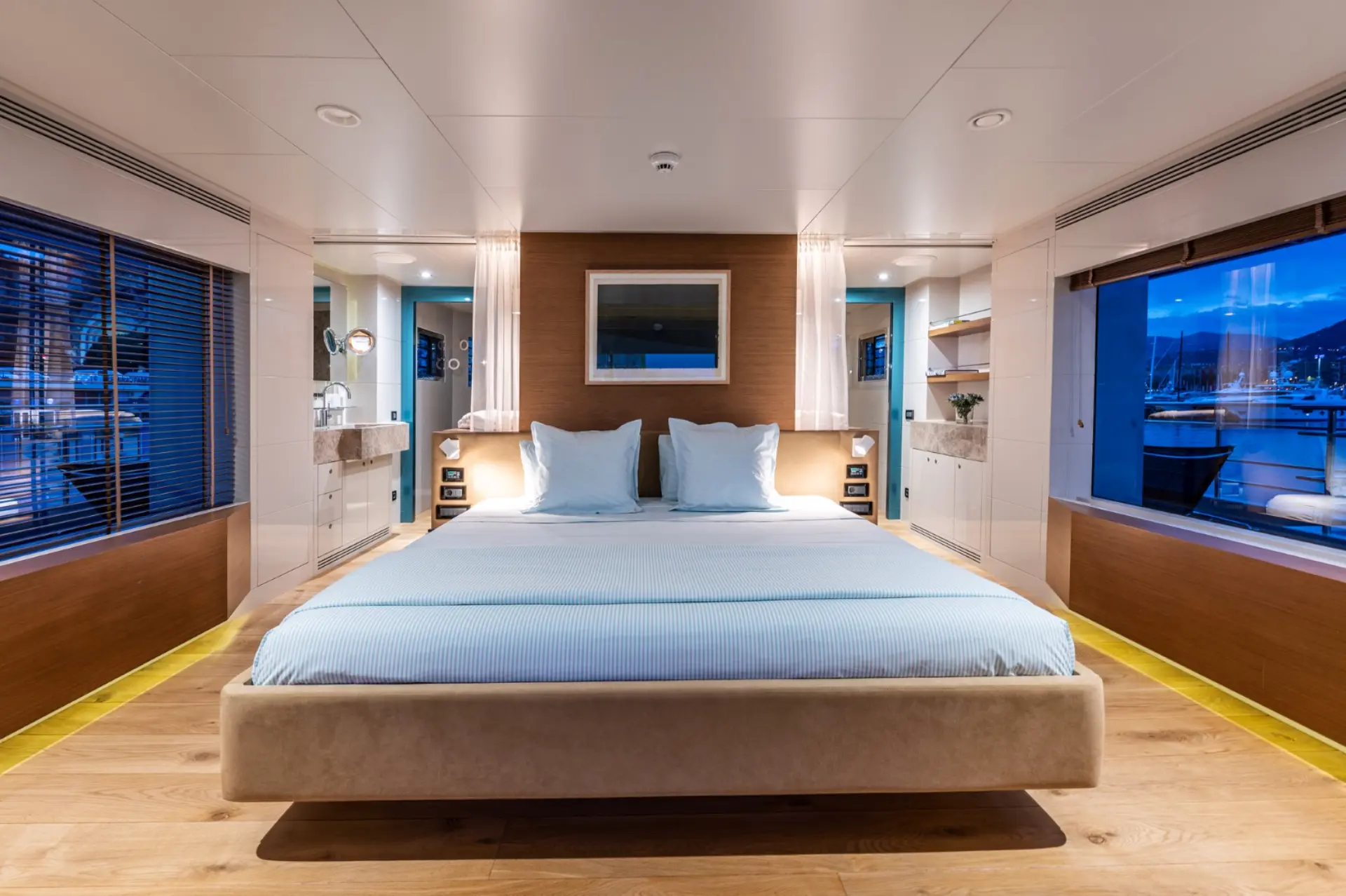 Spacious master cabin with double bed, nighttime marina views through large windows, and bathroom entrance on left side.