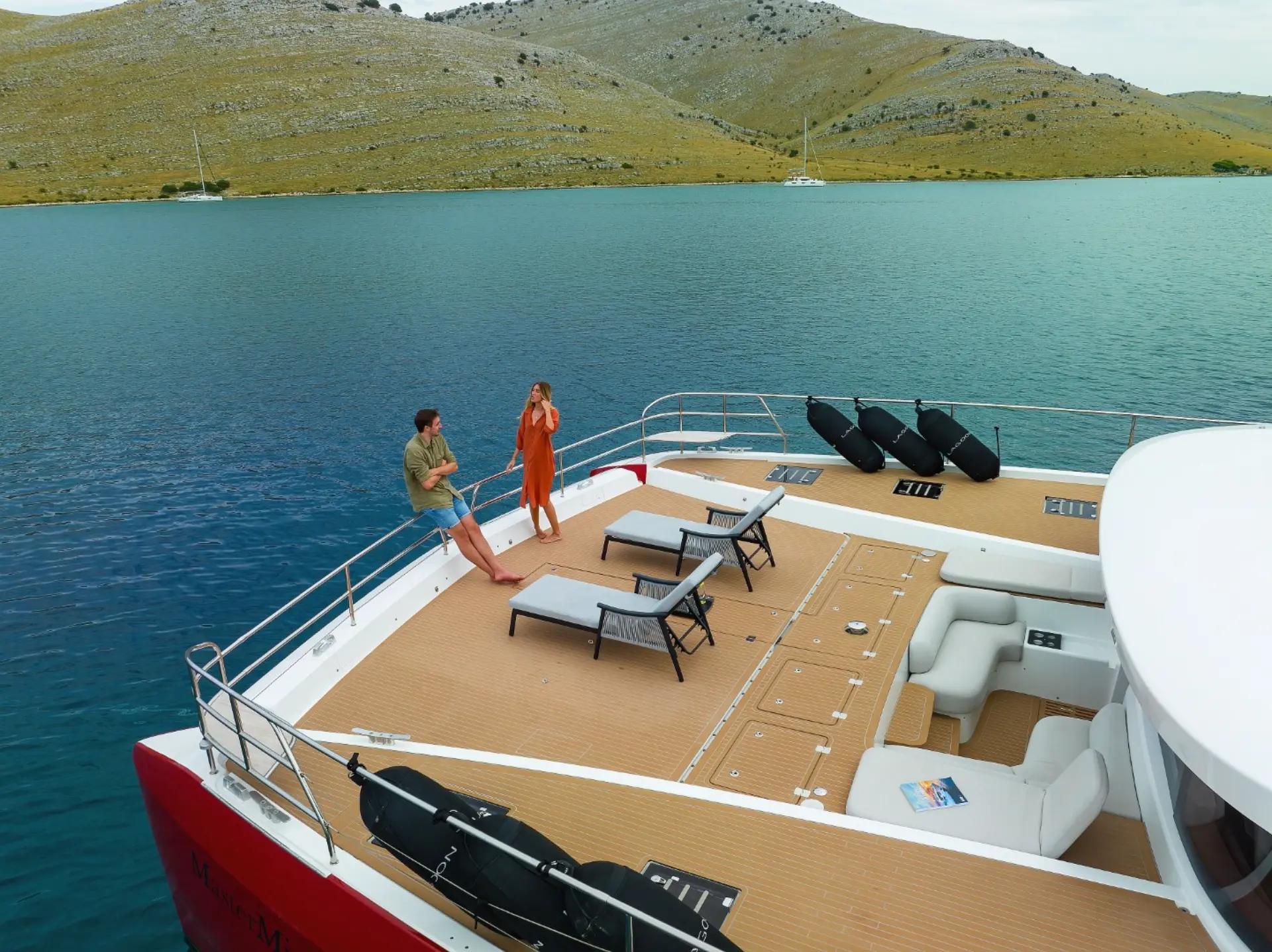 Mastermind – side profile showing foredeck loungers, couple at bow, black fenders and lounge behind.