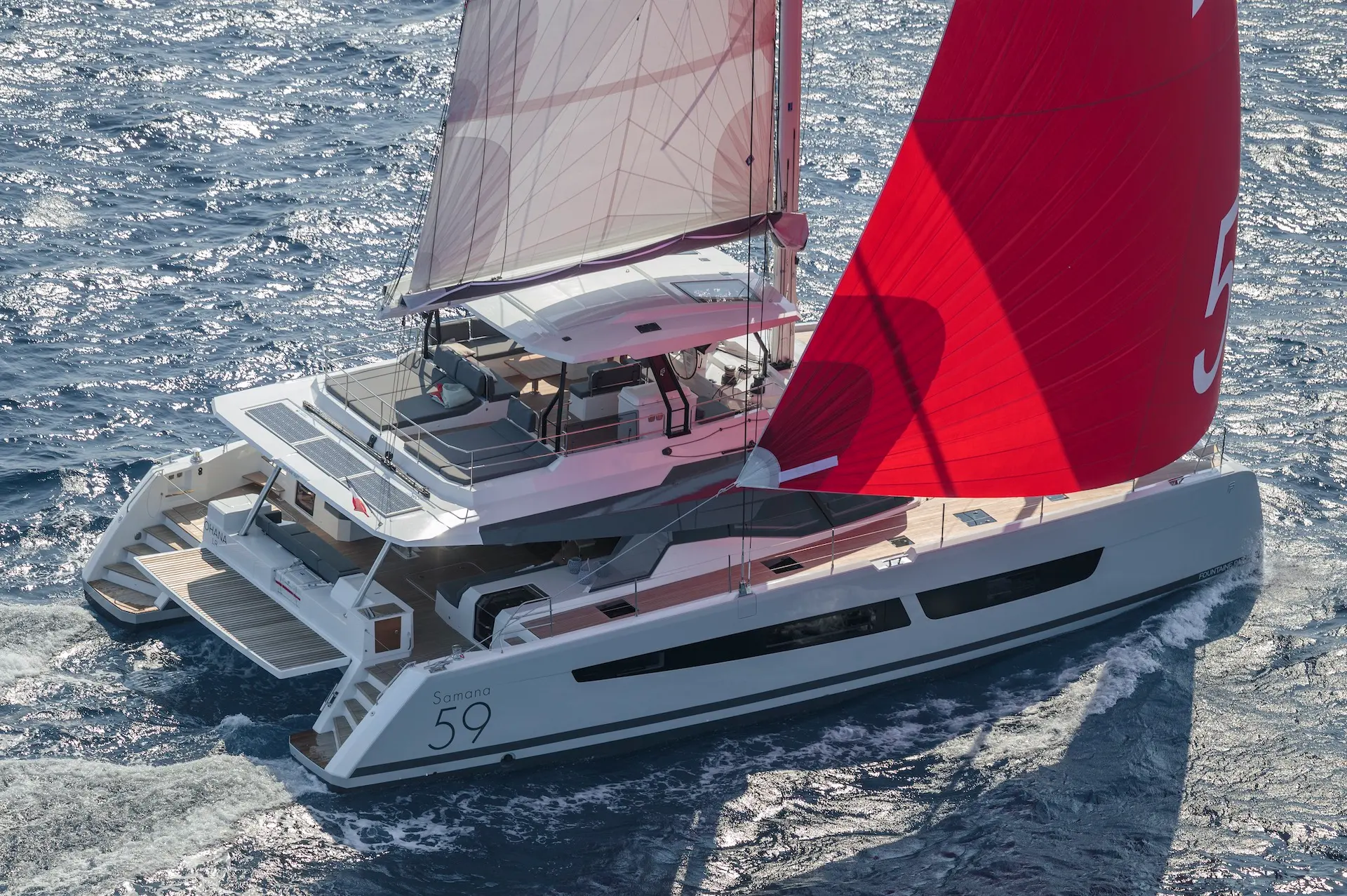 Luxury catamaran Fountaine Pajot Samana 59 – aerial close aft view of yacht sailing with full white and red sails on blue sea.