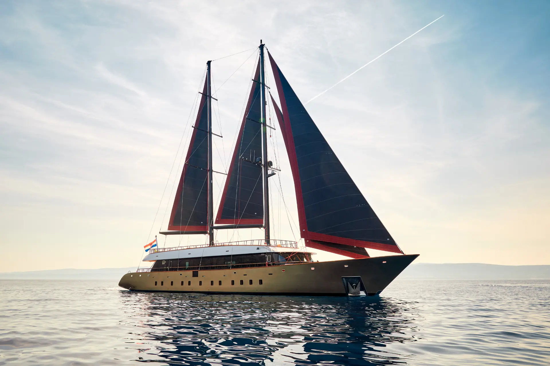 Luxury sailing yacht Anetta - side perspective showing black sails against a bright blue sea backdrop.