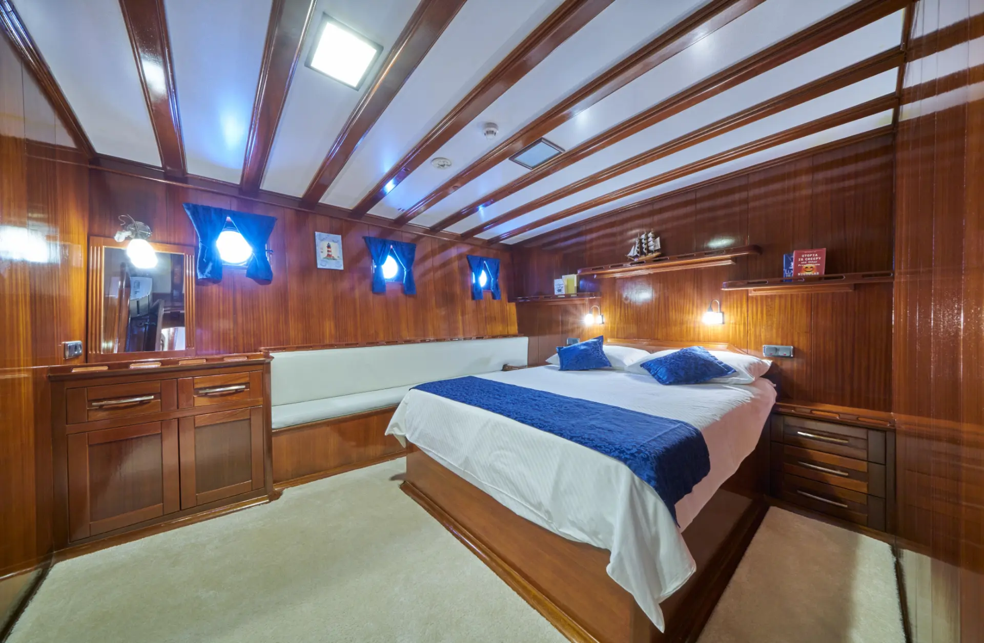 Stella Maris – white and blue bed in double bedroom, comfy seating left, round windows with blue curtains, mirror visible.