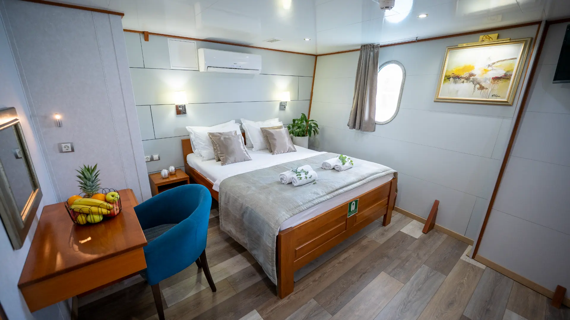 Karizma – double bedroom with comfortable bed, desk topped with fruits, wall art, and oval window.