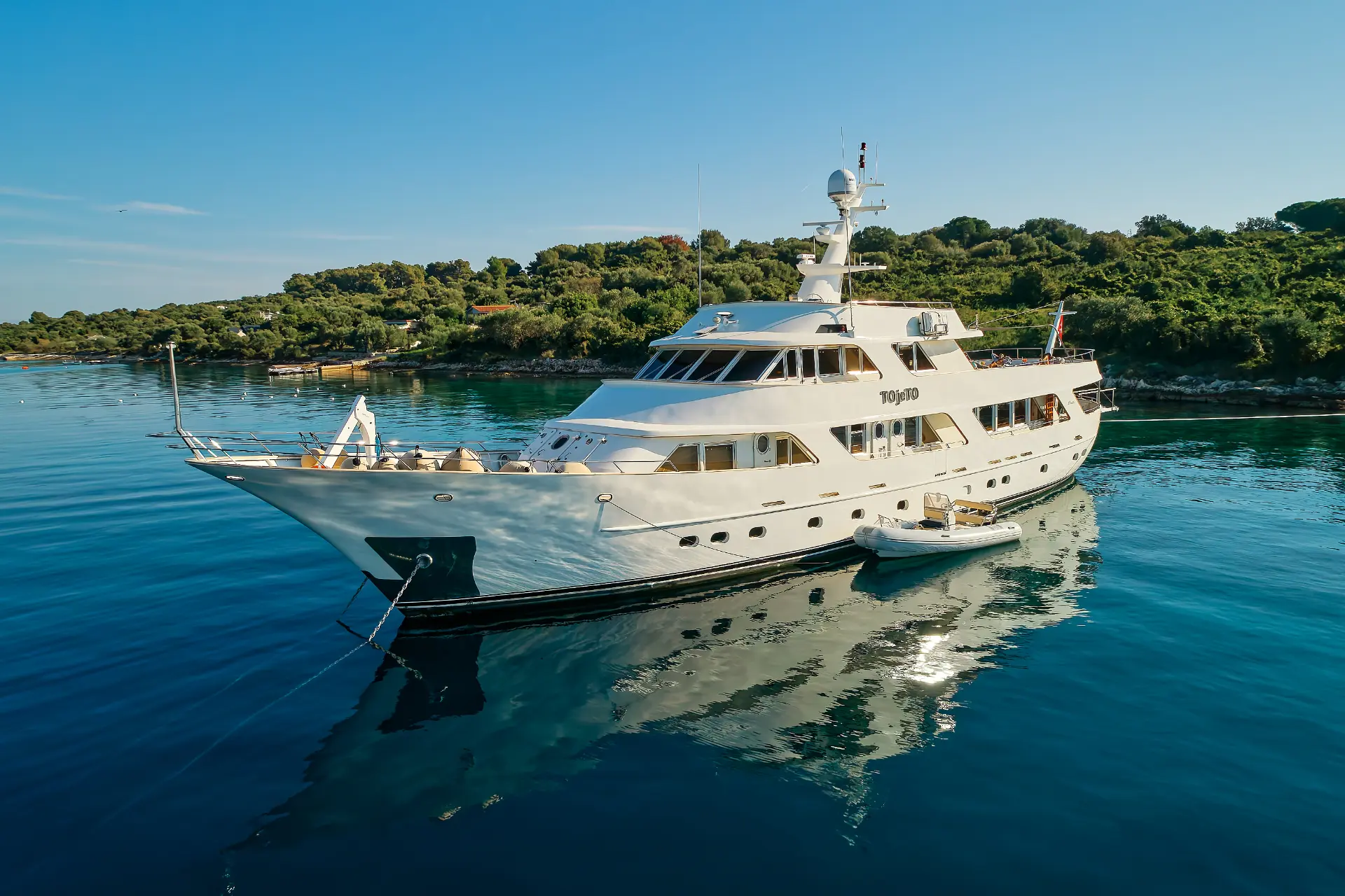 Luxury Motor Yacht To je To – anchored with attached side tender, front diagonal view, lush coastline in background