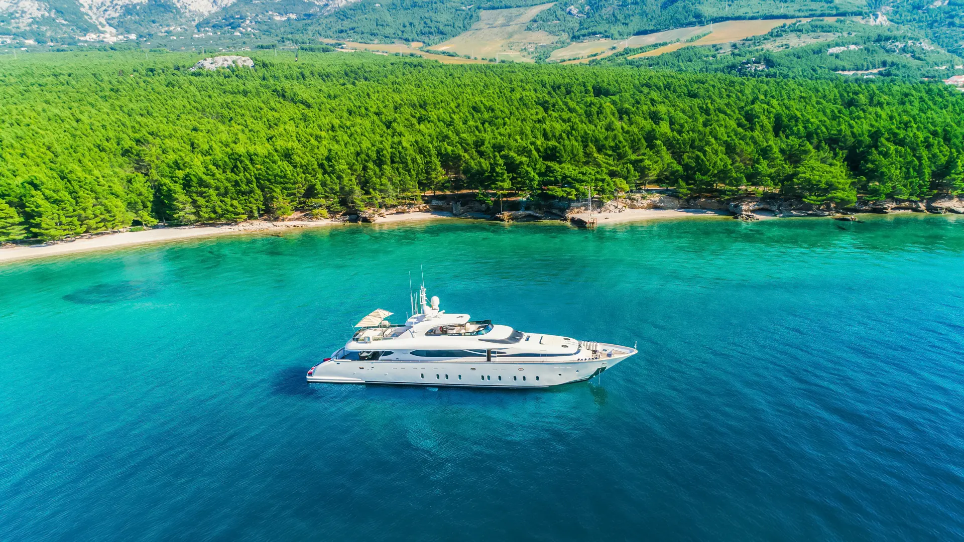 Luxury Motor Yacht Cynthia - aerial side angle, anchored by scenic green coastline under blue skies