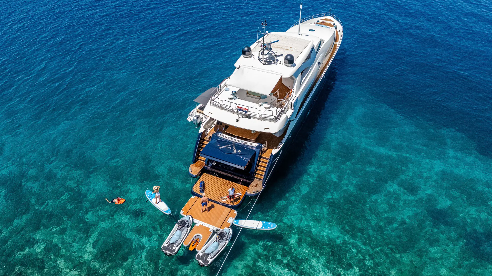 Arago - aft aerial view, anchored with ropes to coast, inflatable platform and water toys in use behind yacht.