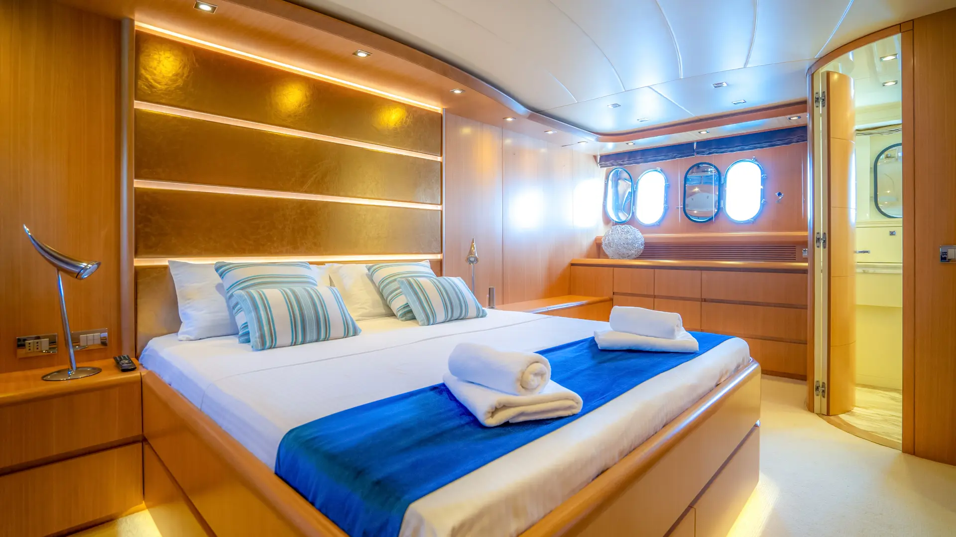 Cynthia – Master cabin with double bed, wooden accents, two oval windows, and bathroom entrance.