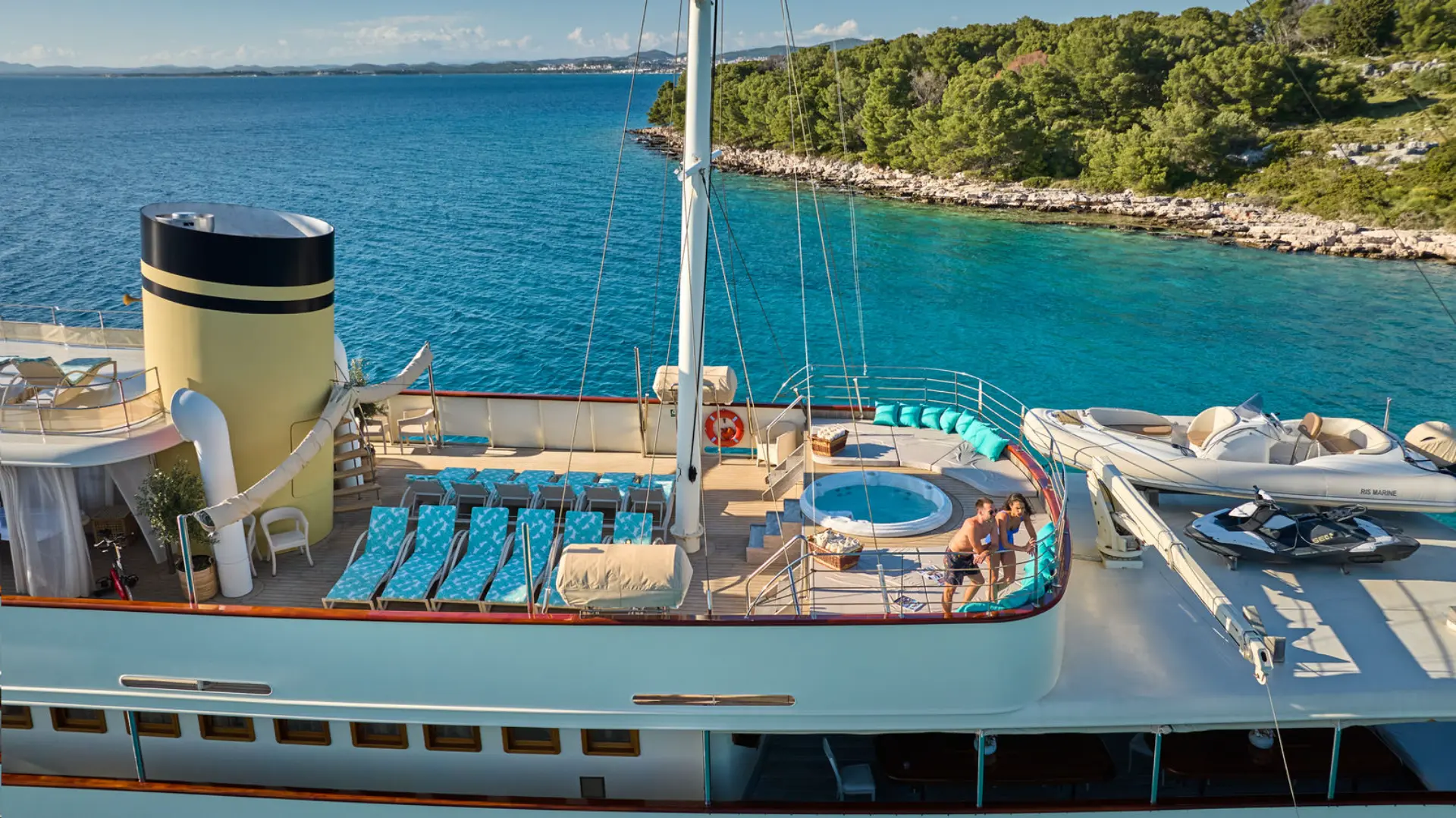 Luxury mini cruiser Casablanca - side aerial shot, upper decks visible, jacuzzi, sunbathing areas, tender, anchored in turquoise bay, lush coastline.