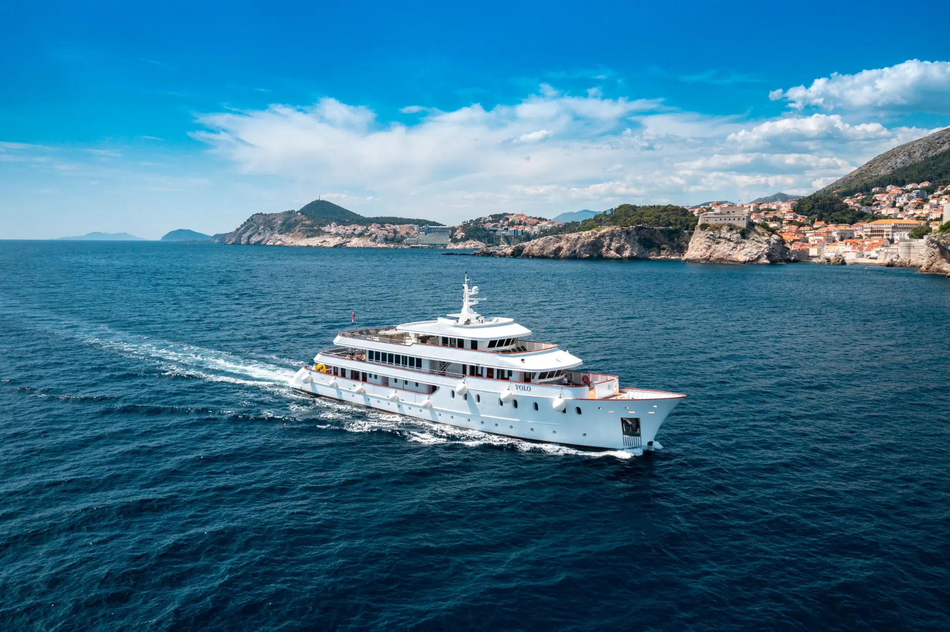 Luxury mini cruiser Yolo - aerial side view , yacht cruising , in back sillouette of Dubrovnik at sunset, gliding past the historic old town walls.