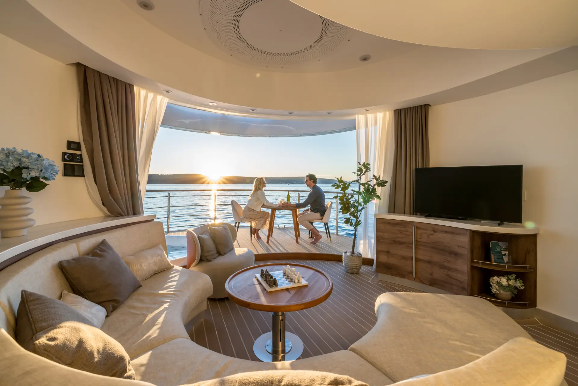 Interior salon view toward bow deck, couple seated on chairs with table between, overlooking a sunset over the sea.