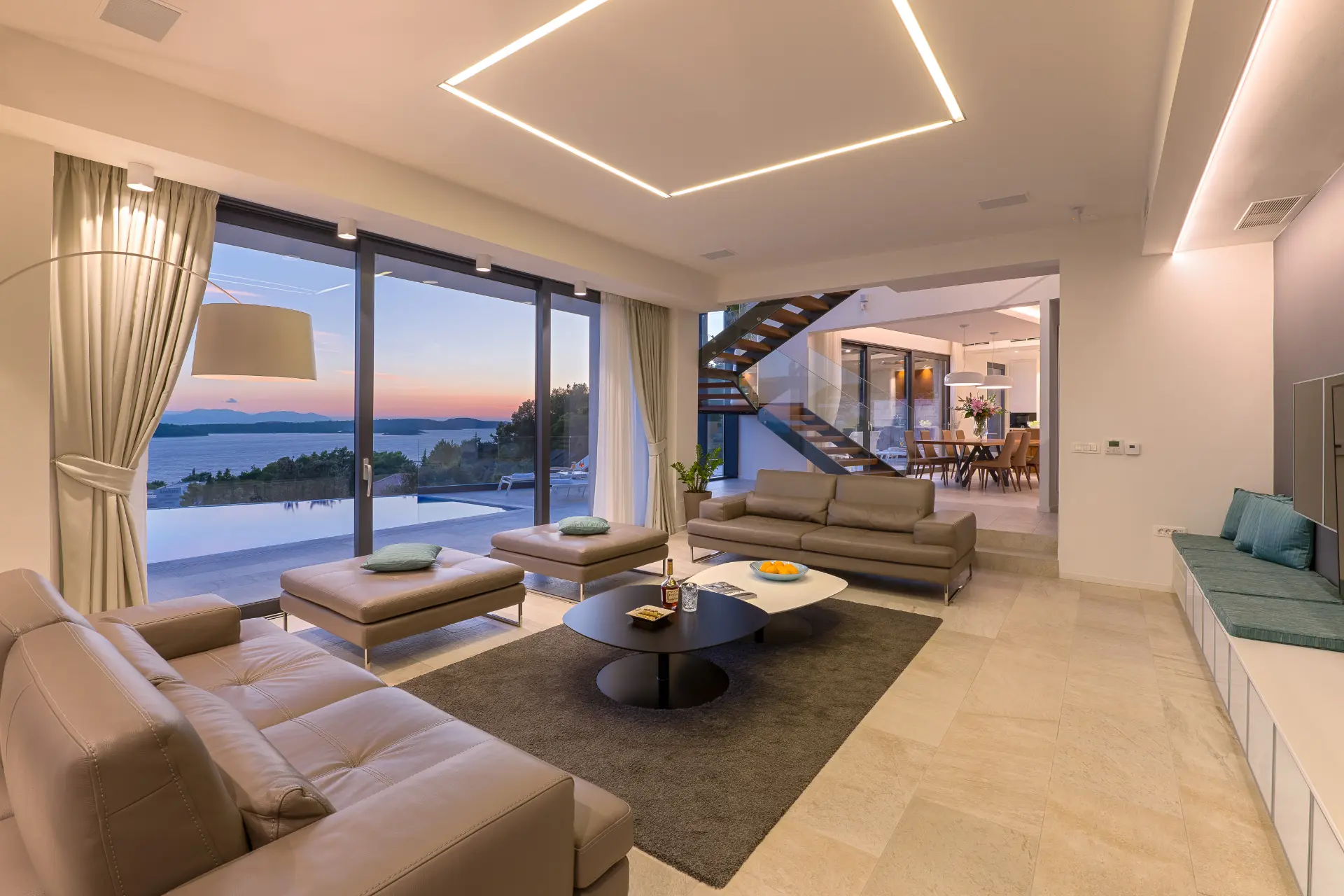 Sunset lit side view of open space living room with seating, glass wall overlooking pool and sea, staircase to first floor, dining and kitchen behind