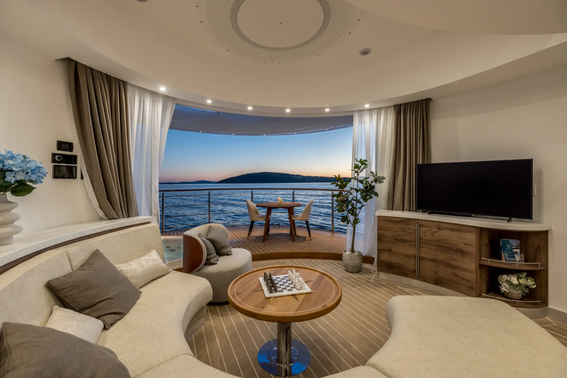 Interior view of Floating House Amor, circular salon with round sofa facing sunset, terrace with table, chairs and jacuzzi visible outside