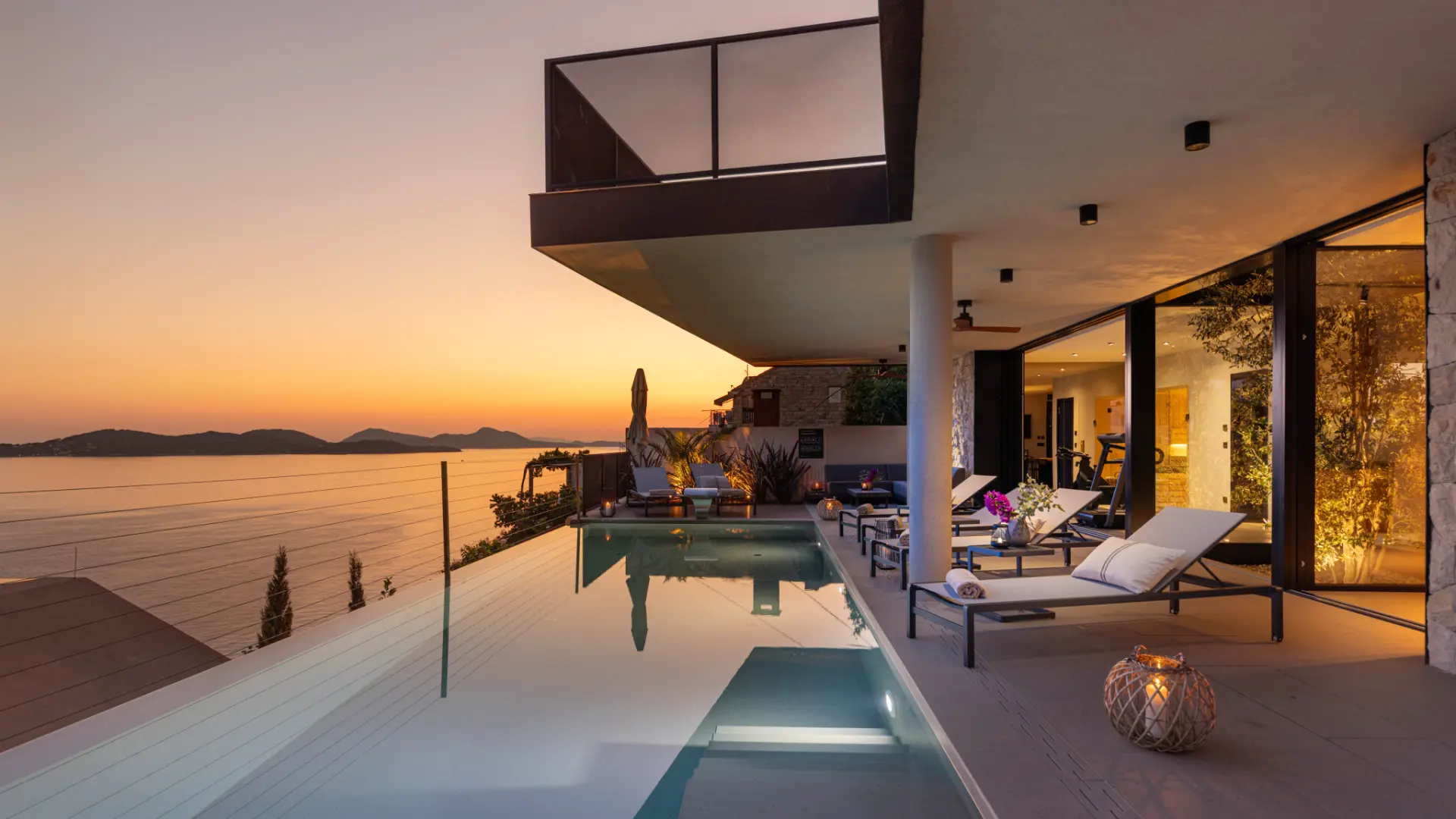 Luxury villas in Dubrovnik - villa Aurea - close exterior view, glowing pool at sunset, sun loungers, glass walls showing indoor fitness area