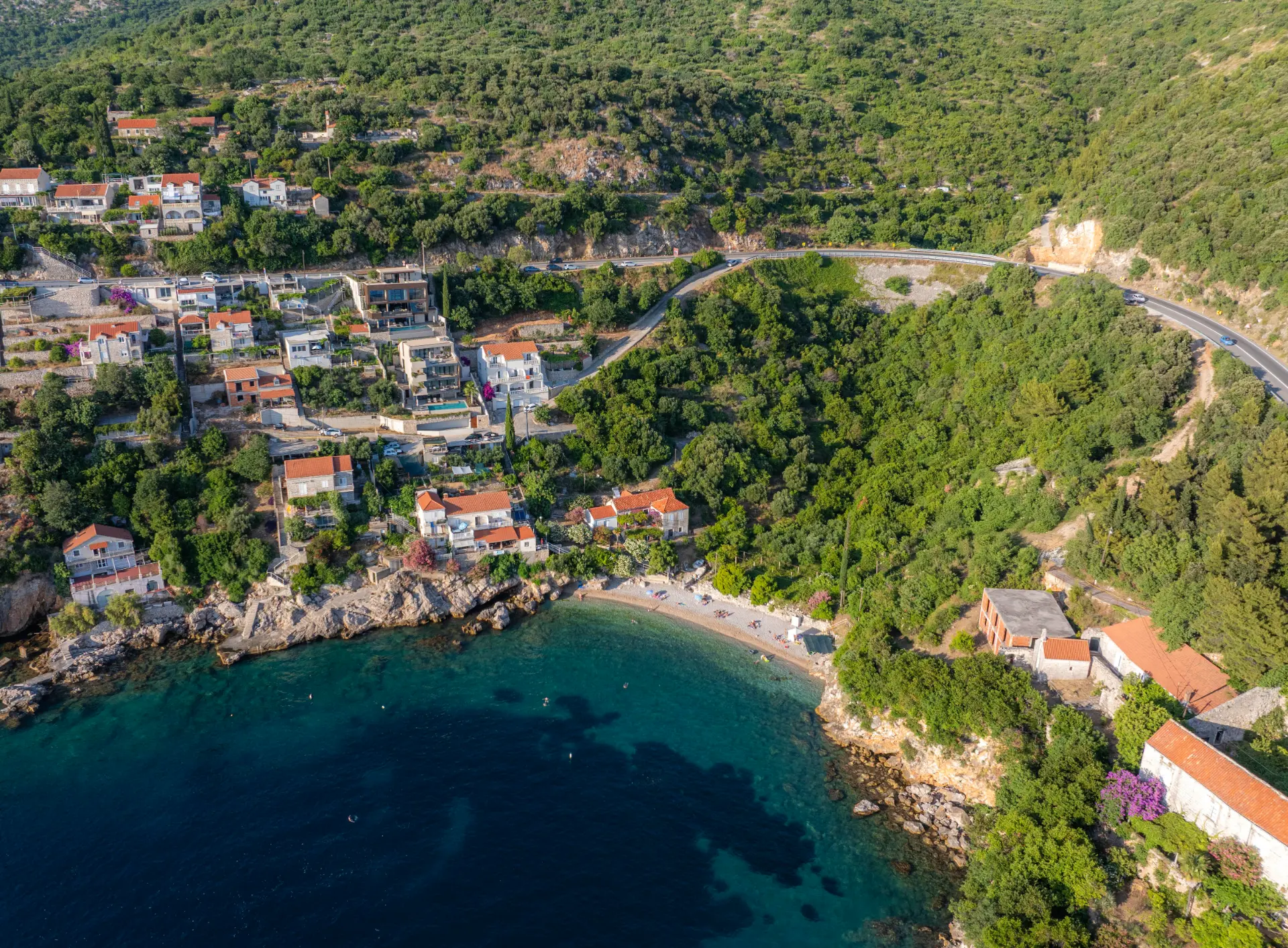 Villa Lumea and Aurea, wide drone perspective of coastal villas set in dense Mediterranean greenery, with a pebble beach and sea on the right