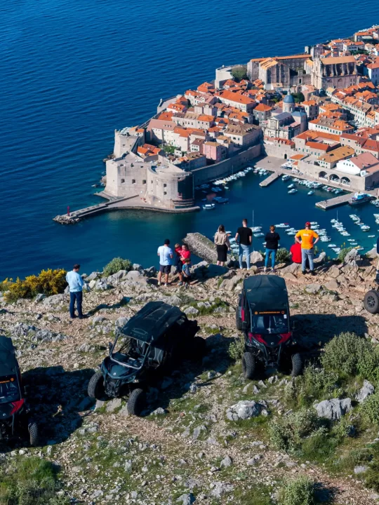 Aerial view of ATVs parked on a hilltop viewpoint above Dubrovnik's Old Town, with the walled city, harbor, and coastline stretching below.