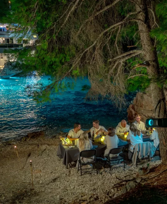 Exclusive Yacht Charter Adriatic - Freedom anchored near pine trees, eight guests seated at table on miniature beach at night