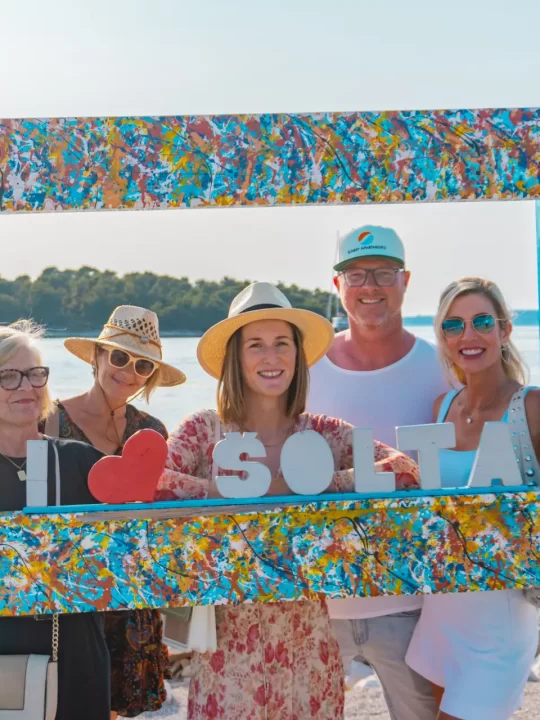 A group of visitors poses behind a colorful mosaic "Šolta" photo frame, with the sea and coastline visible behind them.